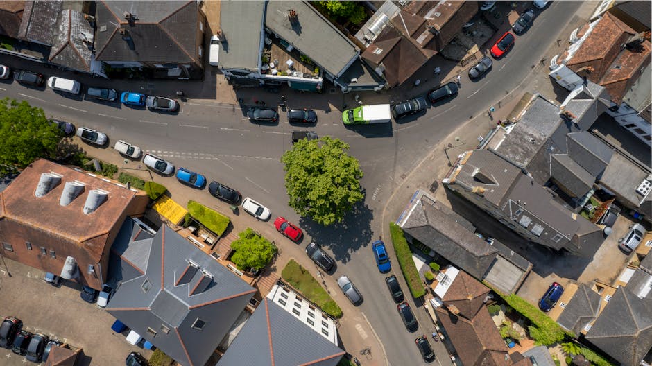 An aerial view of a residential area in Guildford, Surrey, showing a narrow street lined with parked cars on both sides, including sedans, hatchbacks, and vans in various colors such as blue, white, black, and grey. Several houses with pitched roofs, chimneys, and different architectural styles are visible along the street, some with front gardens and driveways. A central tree with lush green foliage provides shade for a few parked vehicles and pedestrians nearby. The surrounding properties include brick and rendered facades, with some featuring small yards or hedges. The scene captures a typical suburban environment during daylight, with natural light casting shadows from the trees and buildings. This image relates to house removals and relocation services offered by Man with Van Surrey, highlighting urban parking considerations and the logistics involved in home furniture transport and packing during local moves.