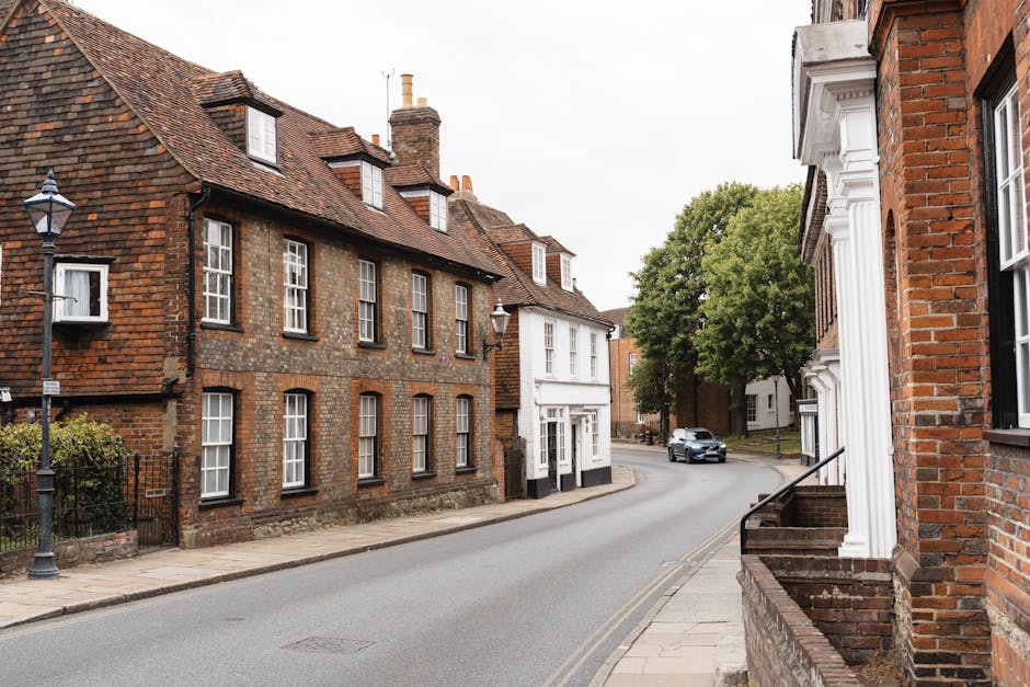 An outdoor street scene in Guildford High Street featuring traditional brick houses with pitched roofs and multiple chimneys. One building has a white facade and large windows, positioned at the corner of a curved road, which is wide enough to accommodate moving vehicles. The street is lined with a sidewalk on both sides, and classic black lampposts are visible along the pavement. In the distance, a silver car is driving along the road, approaching the intersection near a large, leafy green tree. The scene appears during daylight with overcast skies, providing natural, diffuse lighting. This setting illustrates a typical residential area suitable for house removals or furniture transport, with a focus on the street's spaciousness and the architecture of the buildings, aligning with local moving and packing processes as handled by [COMPANY_NAME].