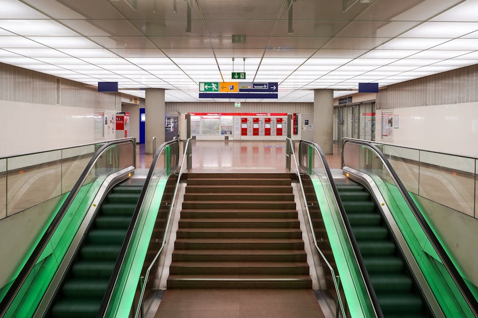 A modern train station platform featuring a multi-level staircase with metal railings and wide steps, constructed from metal and concrete, leading to an elevated walkway illuminated by warm lighting.